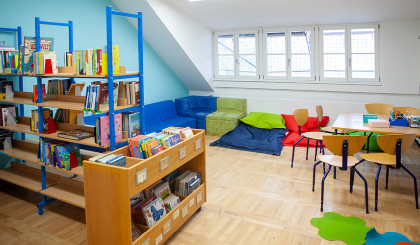 Ein Bild, auf dem ein Raum mit gefüllten Bücherregalen, mehreren gemütlichen Sitzgelegenheiten ein Tisch und mehrere Stühle zu sehen sind.